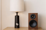 Monitor Audio 8G Silver 50 standmount loudspeaker in walnut finish sat on a wooden table next to a lamp