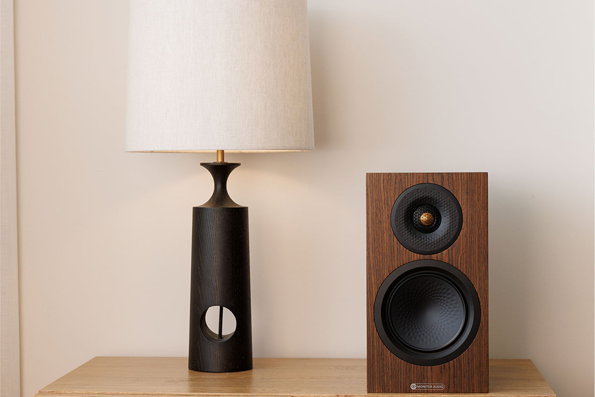 Monitor Audio 8G Silver 50 standmount loudspeaker in walnut finish sat on a wooden table next to a lamp